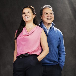 A smiling white woman with long brown hair and a pink top and a smiling gray-haired Asian man with glasses and a blue pullover stand together. 