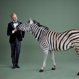 on a green background, an elegant white man in a dark suit leans slightly away from a zebra that is nuzzling him.