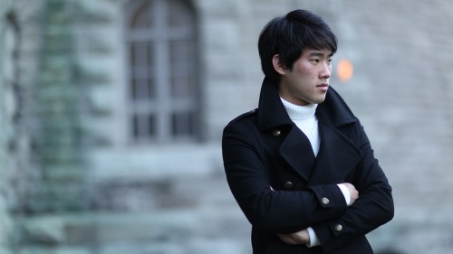 a young east asian man crosses his arms in front as he stands outdoors in front of a stone building. He wears a white turtleneck sweater and a pea coat.