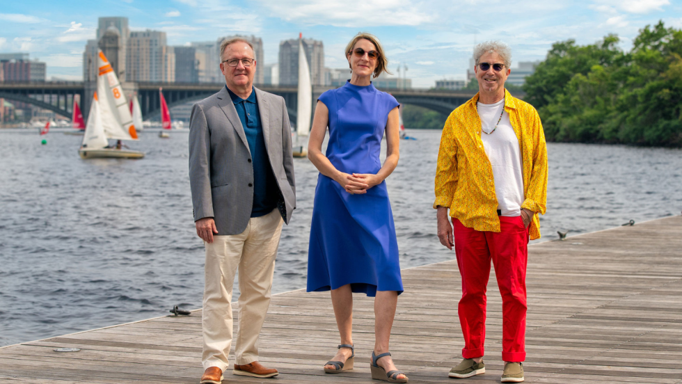 A white man in a blazer and khakis, a white woman in a blue dress, and a white man in a vivid gold shirt and red trousers, smiling on a dock in the Charles River