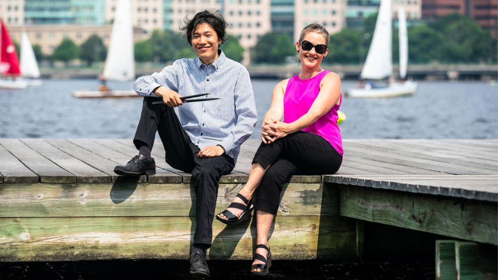 sitting on the edge of a dock in the Charles River, and smiling bemusedly, an East Asian man holding his drumsticks and a white woman leaning back holding one knee